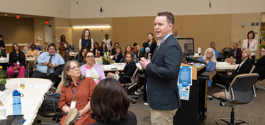 Man stands holding microphone and speaking to people seated at tables in large room