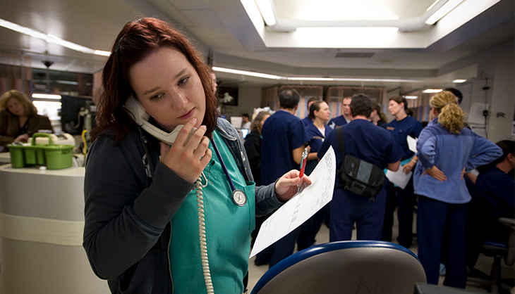 A nurse wearing green scrubs and talking on the phone with a care team standing behind her in the Emergency Department