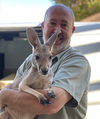 A smiling man with white hair and a beard holds a baby kangaroo in his arms