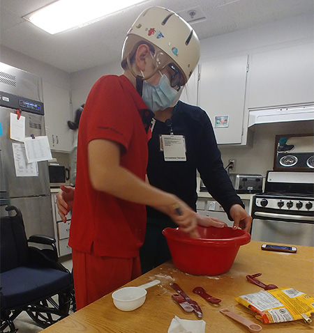 A boy wearing all red with a helmet stirs a red bowl in the hospital kitchen.