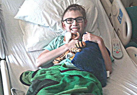 Blonde boy smiles in hospital bed, hugging tiger stuffed animal and blue pillow.