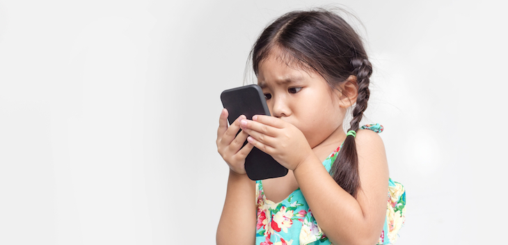 A young girl, with pigtails and wearing a green and yellow sundress, stares closely at a smart phone.