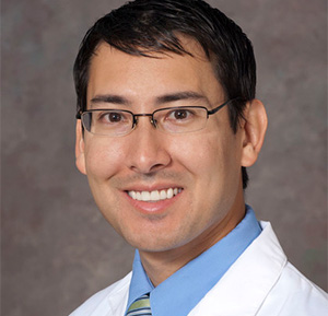 Black-haired man looking into camera with white coat and wearing glasses.
