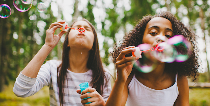 Two girls blow bubbles in an outdoor setting with green trees in the background.