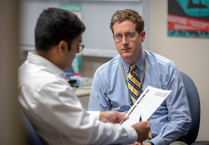 Image of Dr. Stephen Henry wearing a blue long sleeve short and yellow necktie and looking at an individual wearing a white short and holding a paper and pen. Both individuals are wearing eyeglasses.