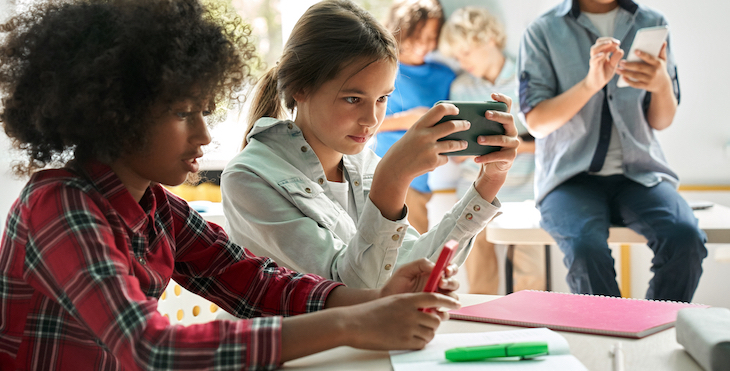 Two girls in a classroom setting are staring at their smartphones with a boy sitting on a desk nearby at his smartphone.