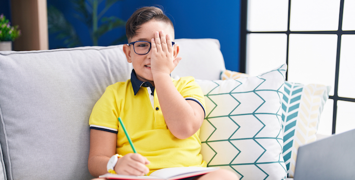 A boy with glasses wearing a yellow shirt is sitting on a couch and holding one hand over one eye.