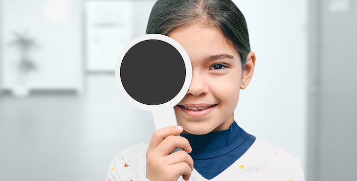 A little girl in a hallway holds a small black paddle over one eye for an eye screening.