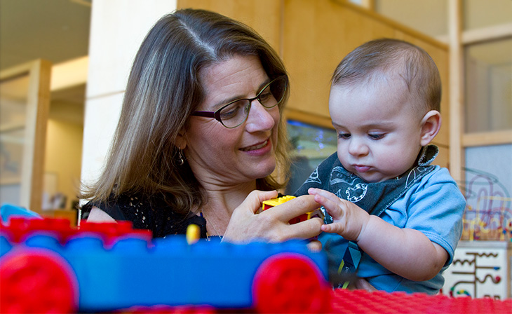 A woman with medium-length brown hair, wearing glasses and a black shirt holds toddler in her arms as they examine a yellow lego-type toy together.