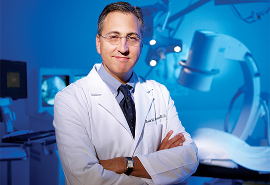 A middle-aged man in glasses and wearing a white physician coat stands with his arms crossed in front of some medical equipment.