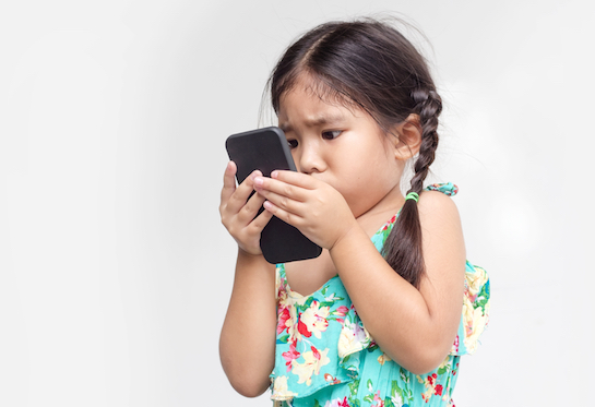 A young girl, with pigtails and wearing a green and yellow sundress, stares closely at a smart phone.