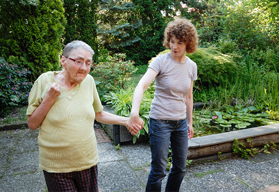 Aging Alzheimer’s patient with short white hair and wearing glasses in a yellow blouse holding hands with a middle-aged woman with curly brown hair