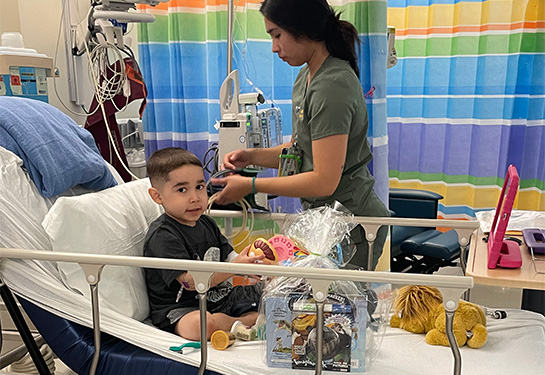 Young boy sits in hospital bed while nurse checks vitals in the background.