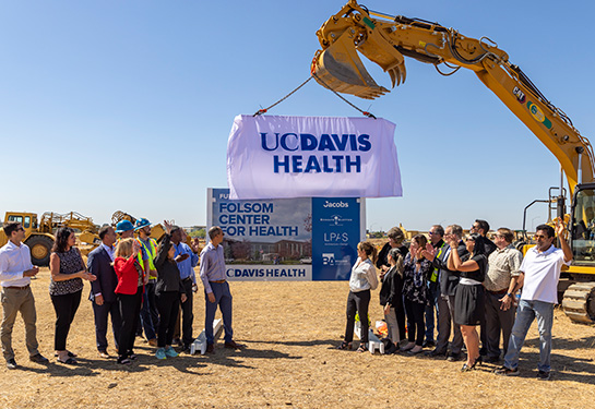 A construction worker uses a large crane to lift up a sign that says “UC Davis Health” to unveil a large sign