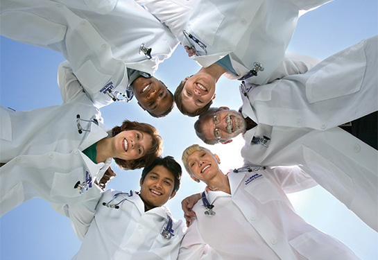 A diverse group of 6 doctors wearing white coats stand with arms around each other in a circle.