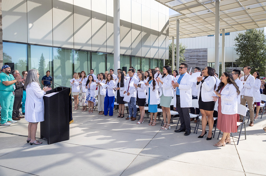  Professor in white coat stands at podium in front of class of students in white coats standing and clapping