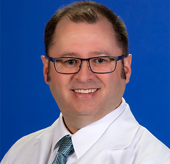 a smiling hematologist Brian Jonas wearing eyeglasses, a white lab coat and white shirt, and a light blue and black checkered necktie. 