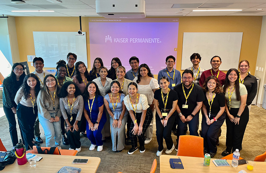 25 teenagers and young adults pose for a photo standing before a blue and white “Kaiser Permanente” logo projected on a video screen