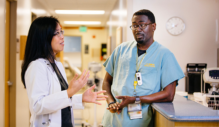 Asian American woman wearing white coat, talking to a African American man in green scrubs