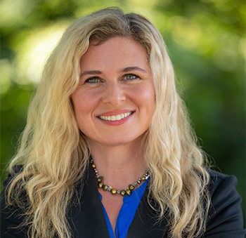 Rachel Whitmer wearing black jacket and blue short pausing in front of the camera with crossed arms. She has blonde hair and green eyes.