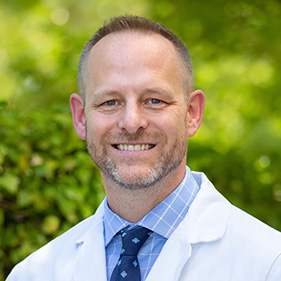 A smiling man with brownish-gray beard and mustache stands in a white doctor’s coat, outside