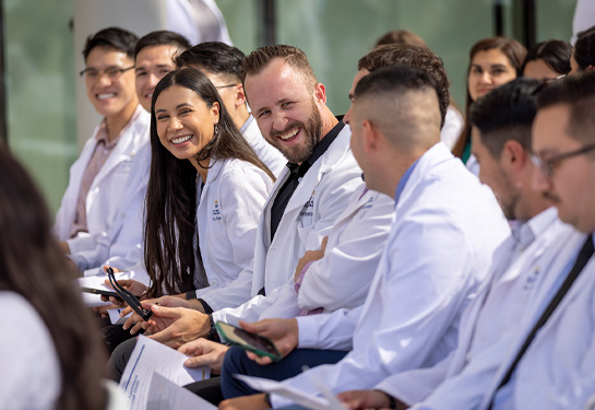 Students wearing white coats sitting in row look at each other smiling