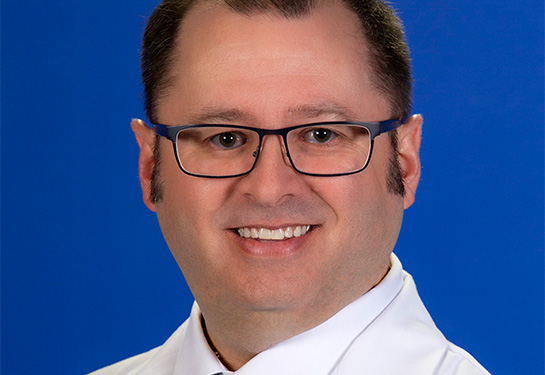 A smiling hematologist Brian Jonas wearing eyeglasses, a white lab coat and white shirt, and a light blue and black checkered necktie.