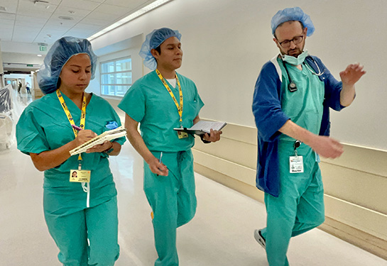A teenage girl and a teenage boy, in green scrubs, take notes as they walk through a hospital hallway led by a doctor in scrubs