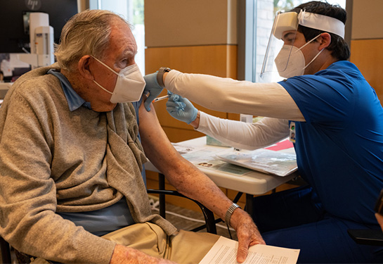 Older man with left arm out sweater getting vaccination in left shoulder
