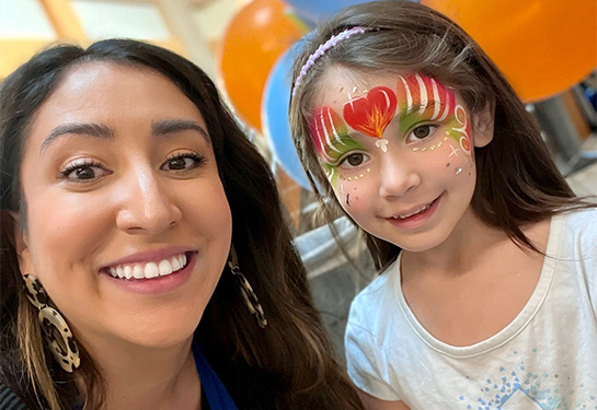 A woman with long, dark brown hair smiles widely next to a young girl whose face has been painted with red and green stripes and a heart. 