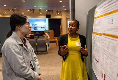 African American woman in yellow dress in front of poster talking to Asian American woman.
