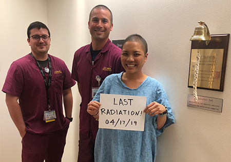 A smiling adult patient in a hospital gown stands next to a gold bell mounted to the wall, with two health care providers in dark red scrubs.