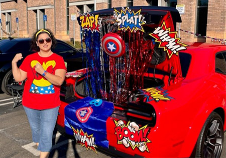 A woman wearing a Wonder Woman shirt, headband, jeans and sunglasses, flexes her arms while standing outside next to a red Dodge Challenger that is decorated in a superhero theme.
