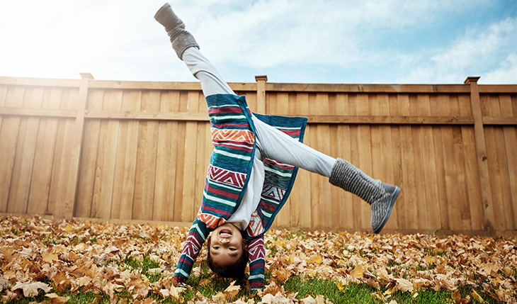 A young girl with long dark hair, wearing a bright multicolored sweater and gray boots does a handstand outside in a yard covered with leaves, in front of a wooden fence. 
