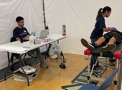 Inside of a medical tent, a boy lies on a table while a female volunteer puts probes on his chest. A man sits with a laptop at a table.