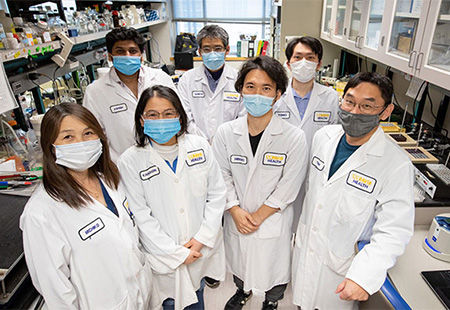 Group of seven people standing together in a lab with white coats and wearing masks