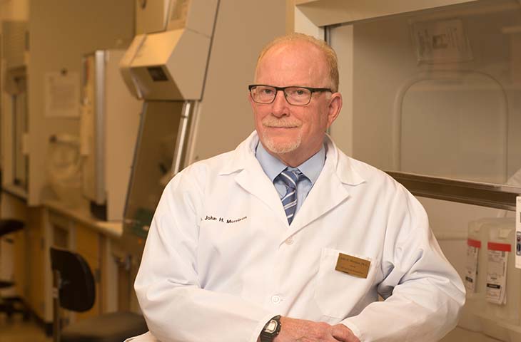 A man with blondish hair, mustache and beard wearing glasses and a white lab coat sits in a research lab facility.