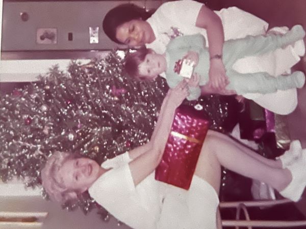 Two women in nursing uniforms sit in front of a Christmas tree. One woman has a small child wearing pajamas in her lap.