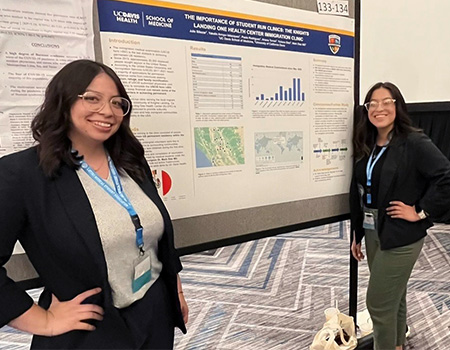 Two young women wearing eyeglasses and dark blazers stand before a white and blue academic poster filled with text and graphics