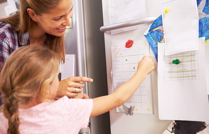 A mom and young daughter put gold stars on an activity chart on a refrigerator together.