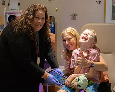 A woman wearing a black cardigan and black pants stands next to a mother holding her young son on her lap while sitting in a medical chair. All are smiling widely and the mother is holding a 