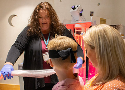 A woman wearing a black long-sleeved cardigan holds a sensory toy made of long plastic strings out to a young boy who's sitting in his mom's lap. 