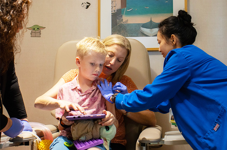 A mother with medium-length blonde hair holds her young son, also blonde, on her lap in a medical chair as a nurse wearing a blue medical coat and blue gloves leans over and cleans the boy’s arm.