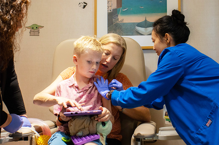 A mother with medium-length blonde hair holds her young son, also blonde, on her lap in a medical chair as a nurse wearing a blue medical coat and blue gloves leans over and cleans the boy's arm.