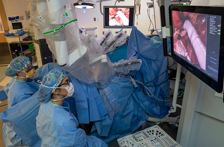 Machine with multiple arms over person laying in bed with two monitors showing surgery