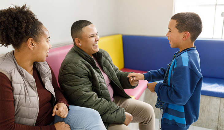 A young boy stands in front of two seated adults in a colorful doctor’s office waiting room. They laugh together. 