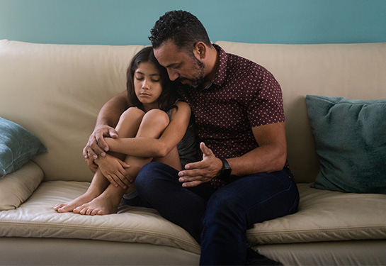 A father and daughter sit on a couch, the child’s knees are up at her chest, and the father is leaning down, consoling her, with his arm around her. 