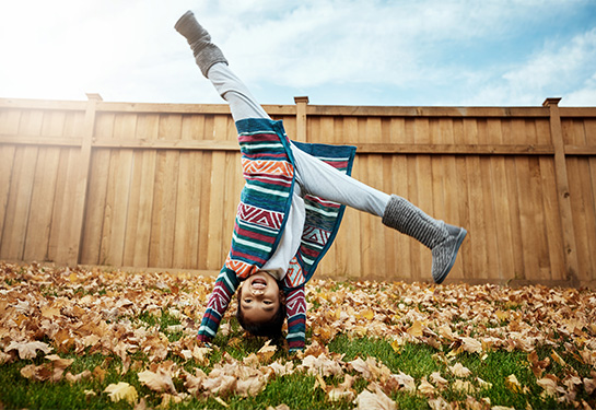 A young girl with long dark hair, wearing a bright multicolored sweater and gray boots does a handstand outside in a yard covered with leaves