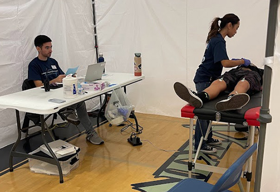   Inside of a medical tent, a boy lies on a table while a female volunteer puts probes on his chest. A man sits with a laptop at a table.