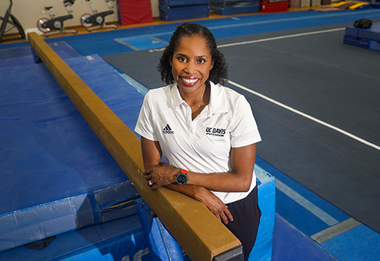 A woman wearing a white polo shirt and watch with a red band stands in a gymnasium leaning on a brown gymnastics balance beam and smiling. 